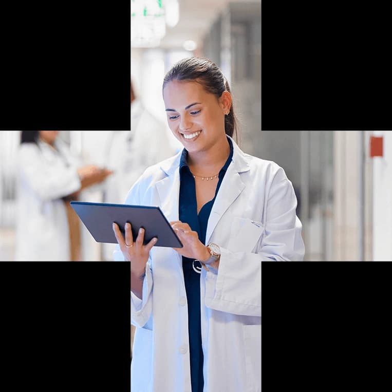 A smiling clinician in a white coat reviewing a tablet in a hospital corridor